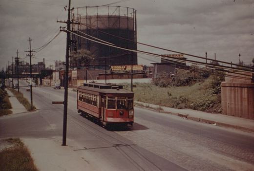 A red and cream wooden streetcar runs down a city street under overhead wires, with a large gas holder and industrial buildings in the background.