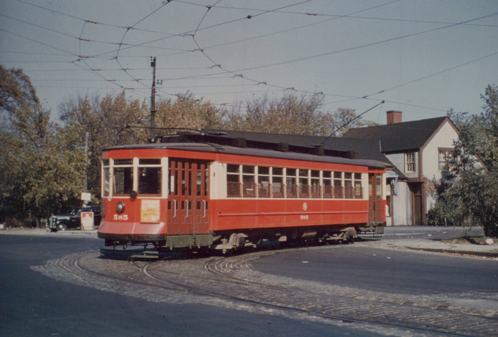A red and cream vintage streetcar, number 585, is turning on streetcar tracks in front of a house, with overhead wires above.