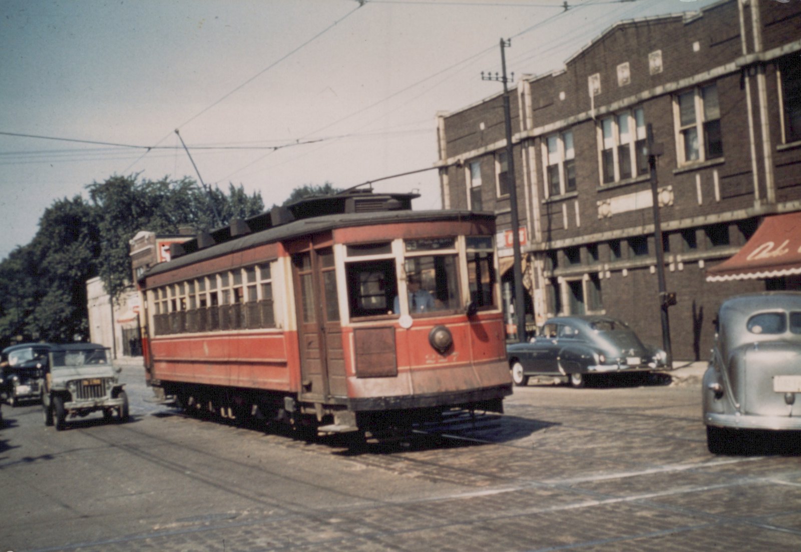 A vintage red streetcar with the number 227 on its front, turning at an intersection with 1940s cars and a brick building in the background.