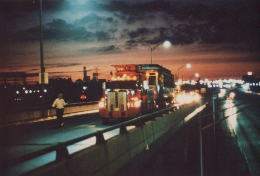 A red, white and blue CTA bus is driving on an elevated highway at dusk, with a person walking on the sidewalk and city lights in the background.