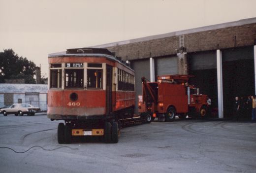 A vintage red streetcar with the number 460 on its rear is being carried on a flatbed tow truck outside a brick building, with a few people standing nearby and a car parked in the background.