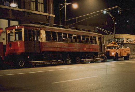 A red and cream vintage streetcar labeled '460' and 'CHICAGO' is being hauled on a flatbed truck down a city street at night, with an orange tow truck and a 'LOUNGE & CATERING' sign visible in the background.
