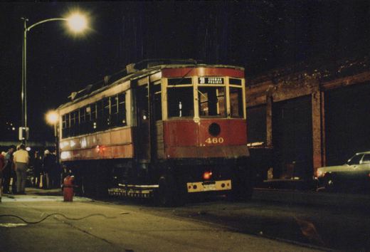 A vintage red and cream streetcar numbered 460 is stopped on a city street at night, with people standing nearby and a car parked on the right.