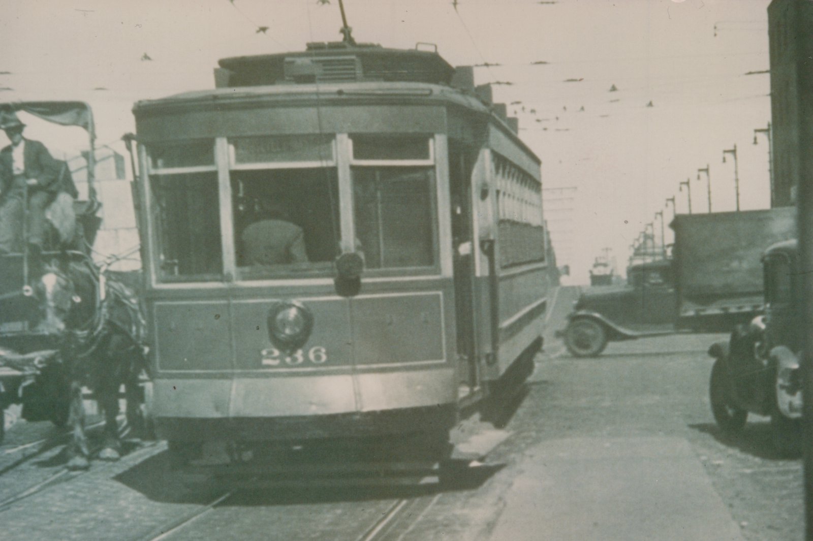 A vintage streetcar numbered 236 travels on a city street alongside a horse-drawn wagon and early automobiles, under overhead wires.