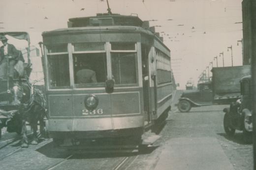 A vintage streetcar numbered 236 travels on a city street alongside a horse-drawn wagon and early automobiles, under overhead wires.
