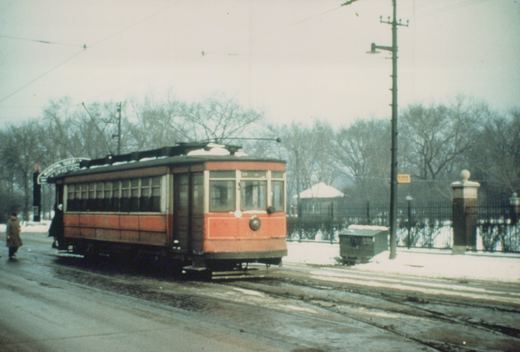 A vintage red streetcar with a destination arch is stopped on a snow-dusted street with bare trees in the background. Two people are near the car.