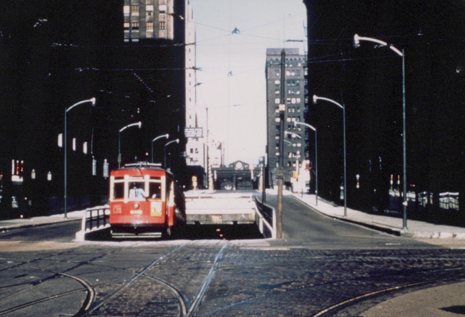 A vintage red streetcar with the number 606 on its front is stopped on a bridge surrounded by tall buildings and streetlights.