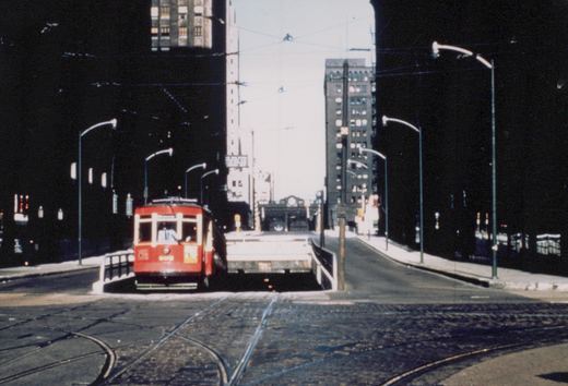 A vintage red streetcar with the number 606 on its front is stopped on a bridge surrounded by tall buildings and streetlights.