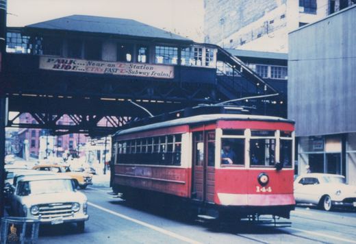 A red and white streetcar numbered 144 runs on tracks in a city street, with an elevated train station and vintage cars in the background.