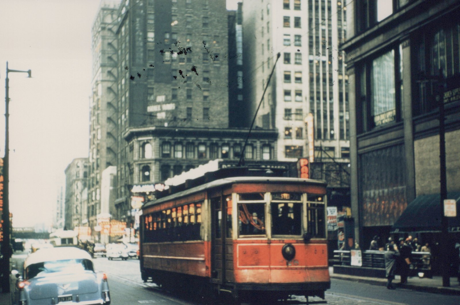A red and cream streetcar with the number 726 and destination Halsted is stopped on a city street, with tall buildings and a 1950s car in the background.