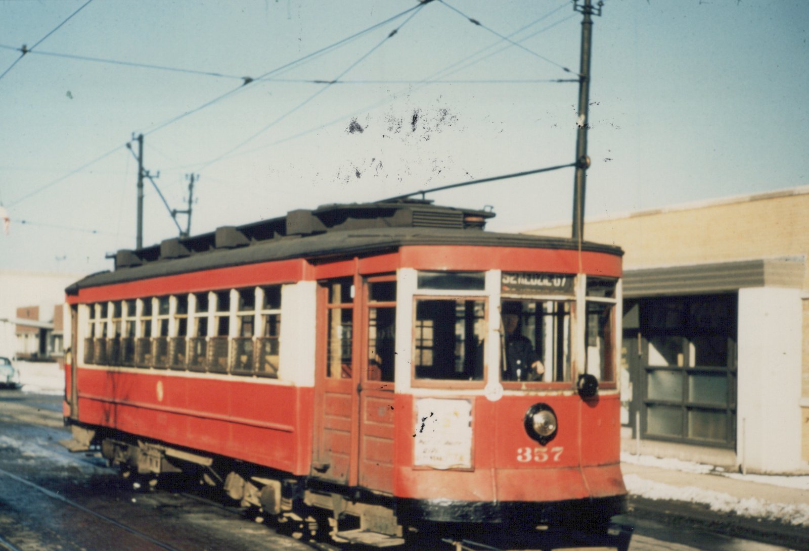 A vintage red streetcar with the number 357 and destination 'California + Roscoe' is shown on a snowy street, with a driver visible in the cab and overhead wires above.