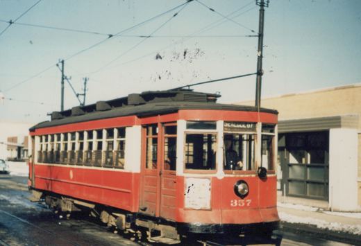 A vintage red streetcar with the number 357 and destination 'California + Roscoe' is shown on a snowy street, with a driver visible in the cab and overhead wires above.