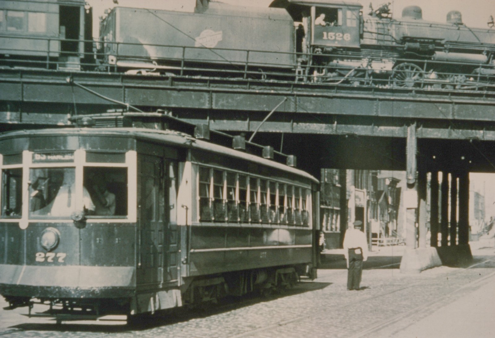 Black and white photo of a streetcar on a cobblestone street with an elevated railroad track above it, carrying a steam locomotive.