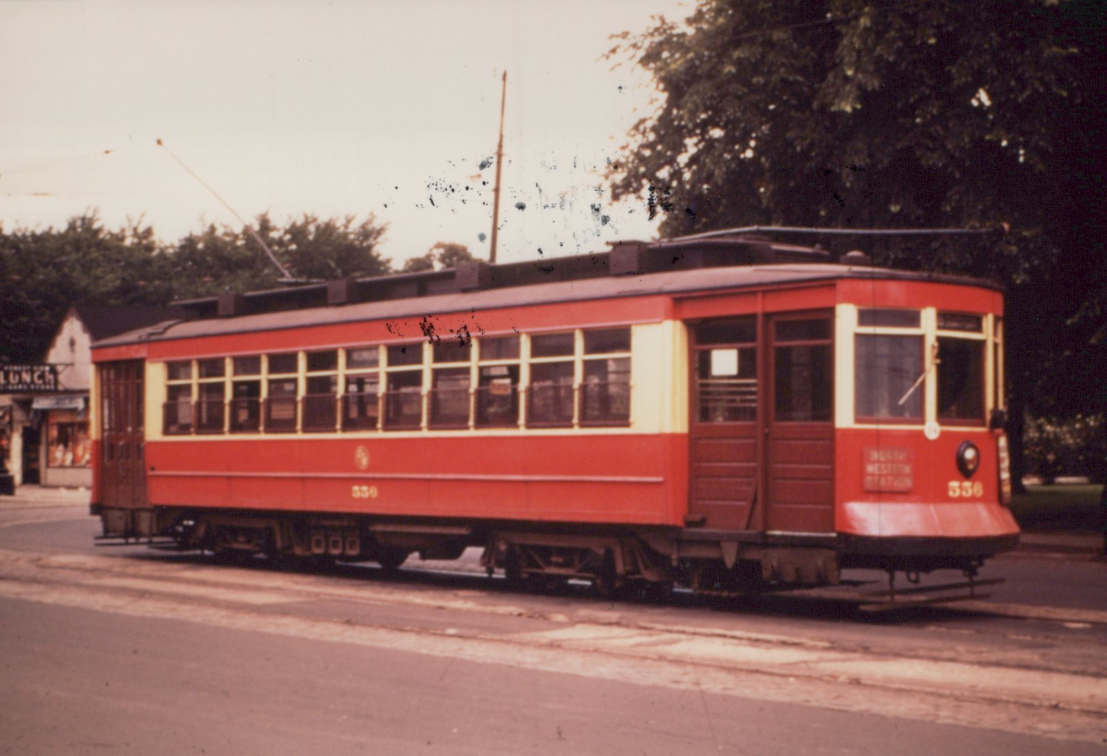 A vintage red and cream streetcar, number 556, is parked on a street with a sign for 'NORTH WESTERN STATION' on its front, a trolley pole reaching to overhead wires, and a 'LUNCH' sign on a building in the background.