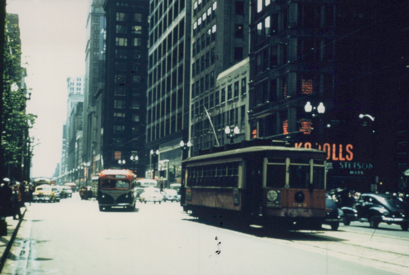 A vintage streetcar and a bus on a wide Chicago street lined with tall buildings and 1940s cars.