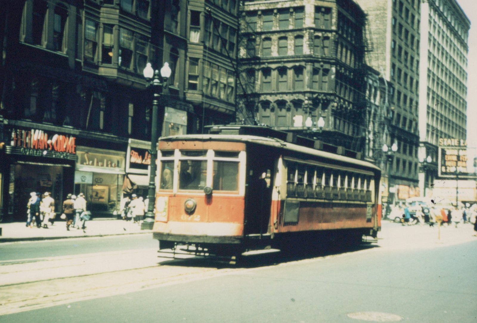 A red and cream streetcar with the number 672 on its front navigates a sunny city street, with pedestrians on the sidewalk and storefronts in the background.