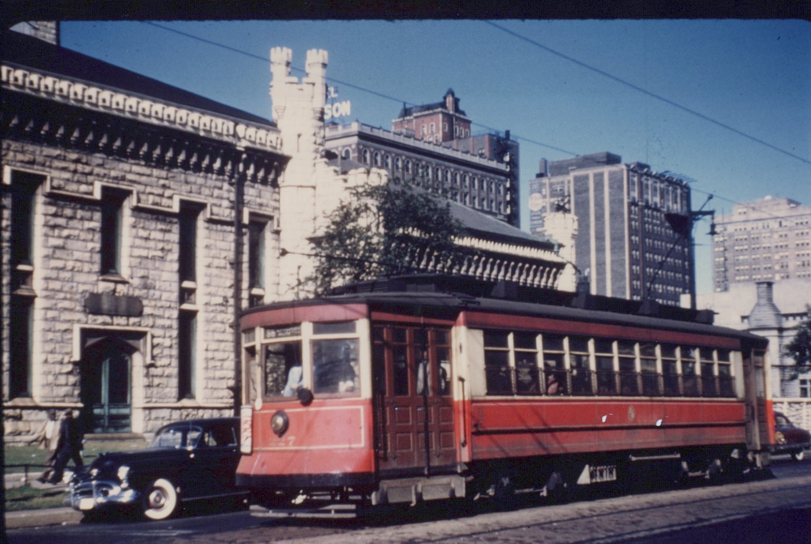 A red and cream vintage streetcar with the number 7 on its front, running on city streets with old buildings and a 1940s car in the background.