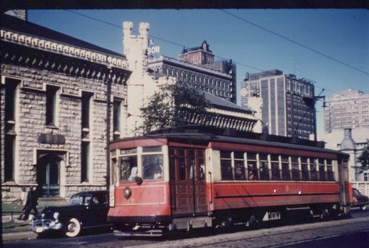 A red and cream vintage streetcar with the number 7 on its front, running on city streets with old buildings and a 1940s car in the background.