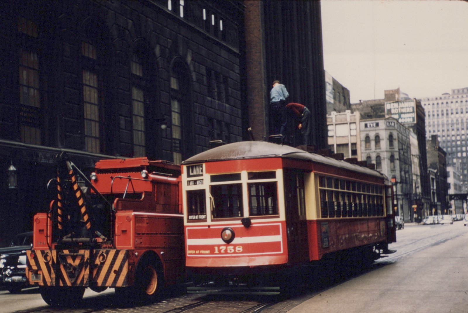 A red and cream streetcar marked 'LAKE' and number 1758 is stopped on a city street with a red work vehicle beside it and two men on its roof, with tall buildings in the background.
