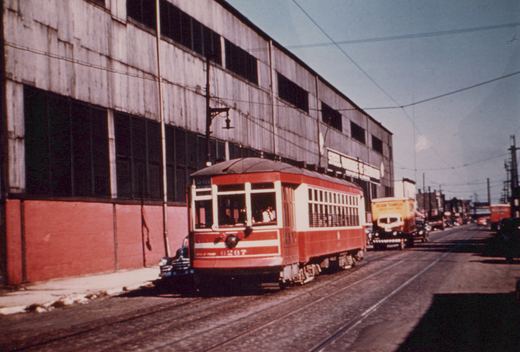 A red and cream streetcar with the number 6267 on its front is parked on a city street next to a large industrial building with a sign reading 'Steel Warehouse & Terminal.' A yellow truck and other vehicles are visible in the background.