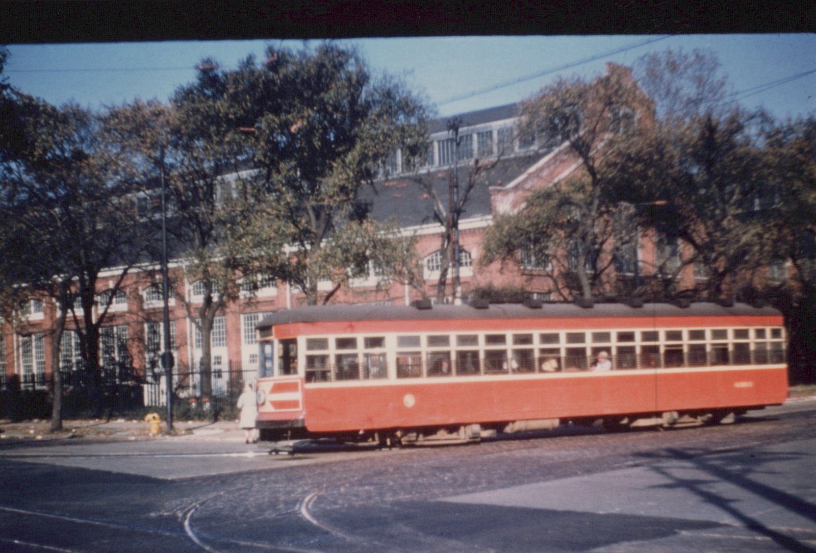 A red and cream streetcar with the number 3111 turns on a street corner, with a brick building and trees in the background.