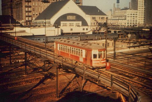 A red and white interurban car with the number 2001 on an elevated steel trestle, with a boxcar train and city buildings behind it.