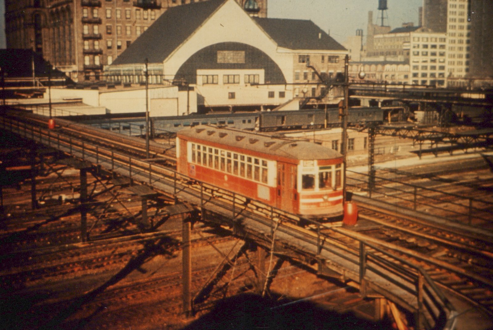 A red and white interurban car with the number 2001 on an elevated steel trestle, with a boxcar train and city buildings behind it.