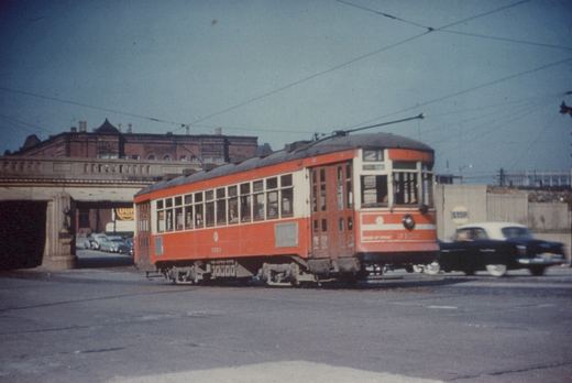 A red and cream streetcar numbered 3153 on route 21 is shown on a city street with an underpass in the background and a 1950s car driving past.