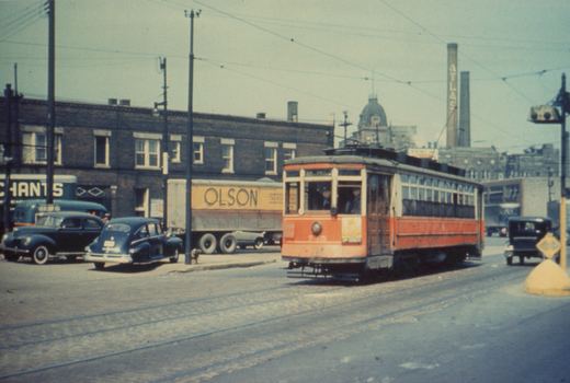 A red and cream streetcar with the number 7-45 on its front is stopped on a city street with several vintage cars and a truck, under an overcast sky.