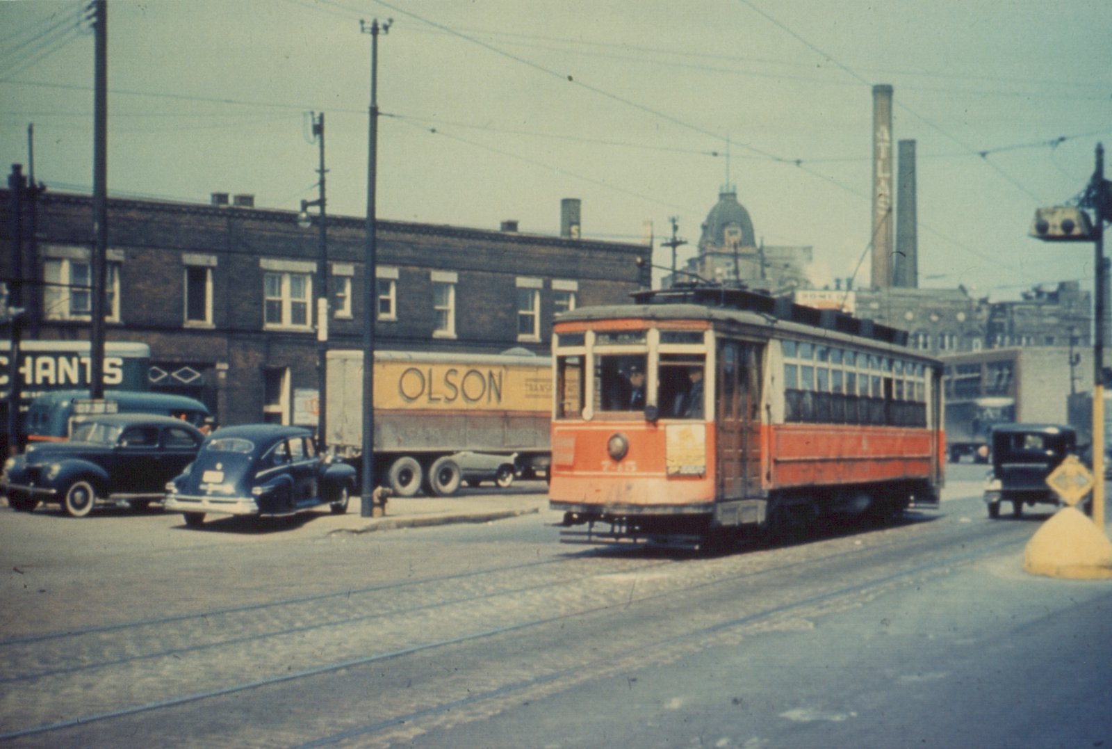 A red and cream streetcar with the number 7-45 on its front is stopped on a city street with several vintage cars and a truck, under an overcast sky.