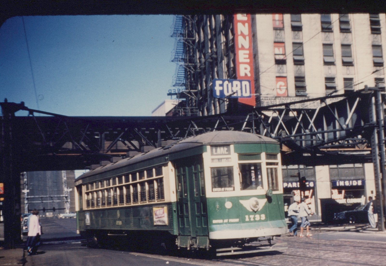 A green and cream Chicago Surface Lines streetcar numbered 1733 is stopped on a city street under an elevated structure, with pedestrians nearby and storefronts in the background.