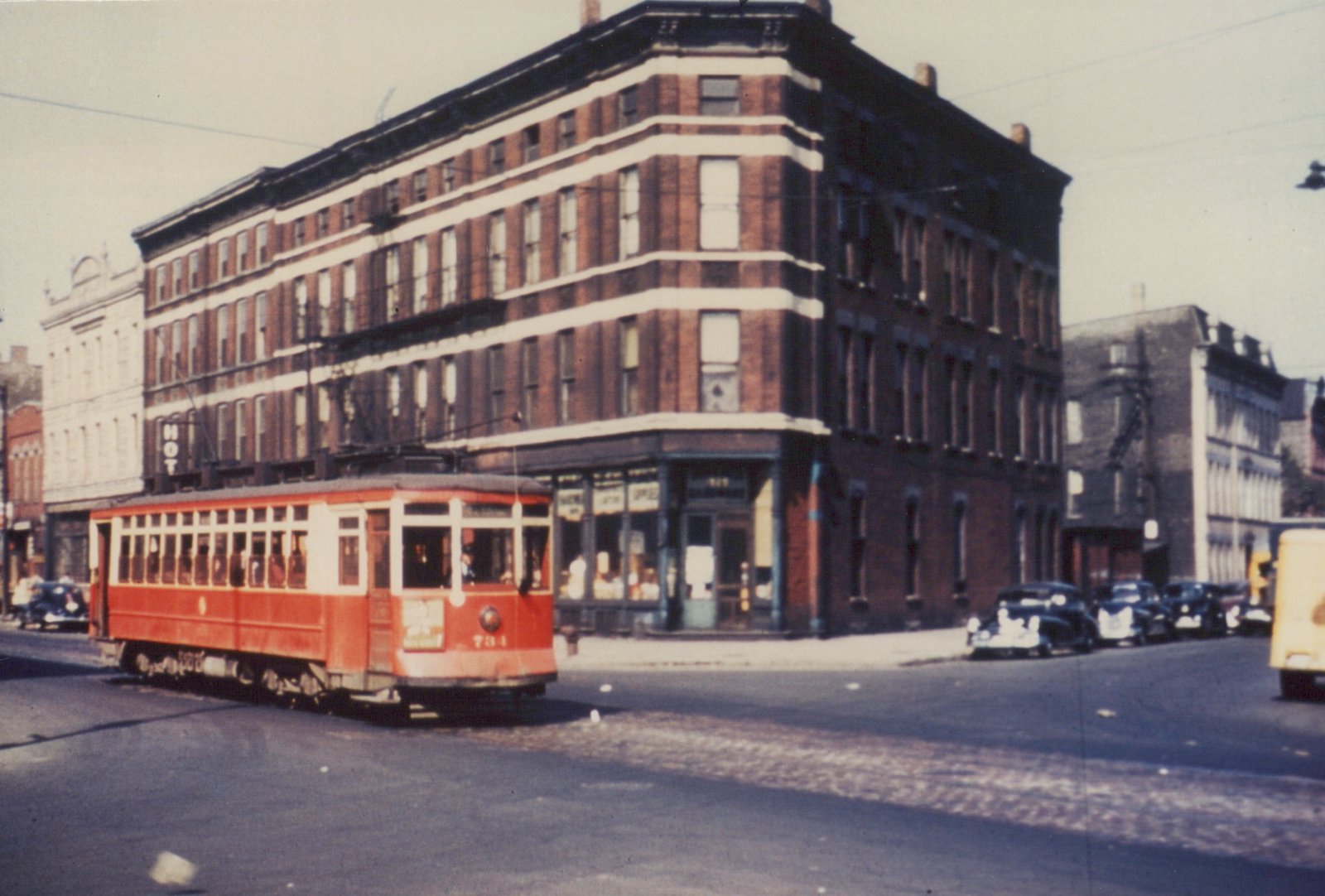 A vintage red streetcar, number 734, is turning a corner on a city street lined with brick buildings and parked cars from the 1930s.