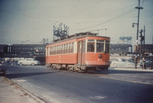 A red and cream streetcar numbered 507 is stopped at an intersection with an elevated train bridge in the background, under a clear sky, with several vintage automobiles and a person standing on the sidewalk.