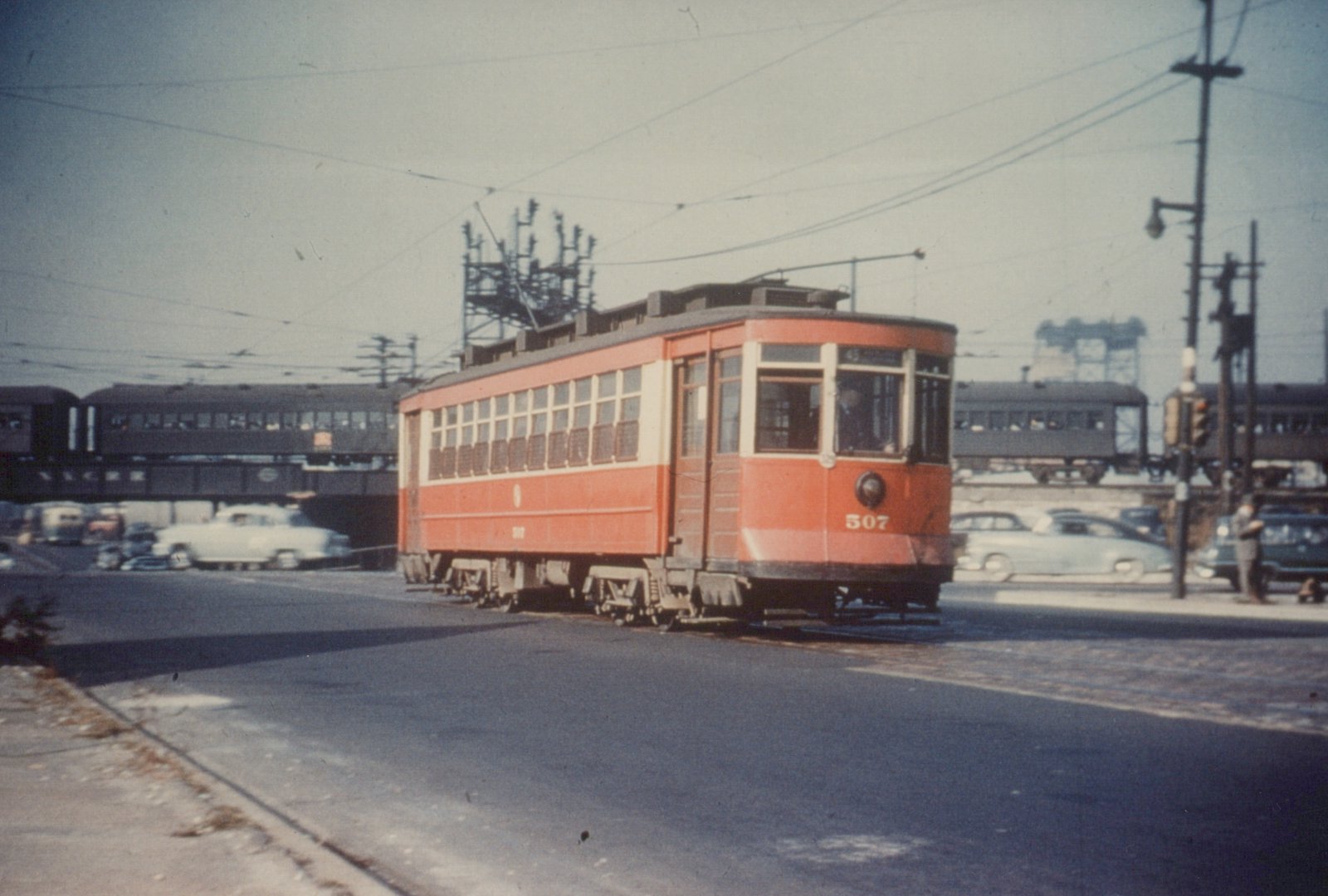 A red and cream streetcar numbered 507 is stopped at an intersection with an elevated train bridge in the background, under a clear sky, with several vintage automobiles and a person standing on the sidewalk.