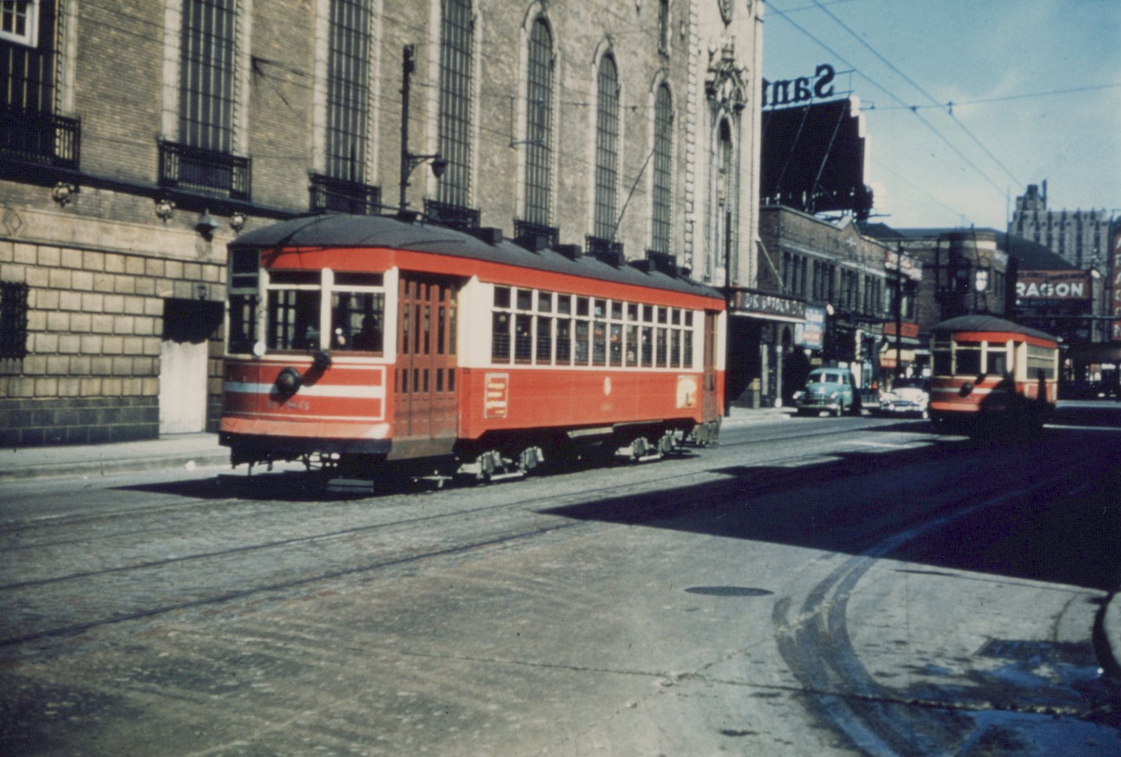 A vintage color photograph of two red Chicago streetcars on a street with a large brick building and billboards in the background.