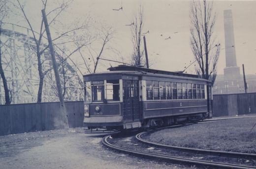 A vintage black-and-white photo of a dark-colored electric streetcar numbered 989 on curved tracks, with bare trees, a wooden fence, and a roller coaster structure behind it.