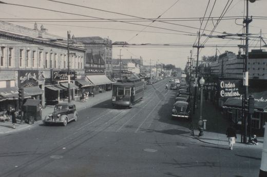 A black and white photo of a vintage streetcar on a city street, surrounded by parked cars and people on the sidewalks.