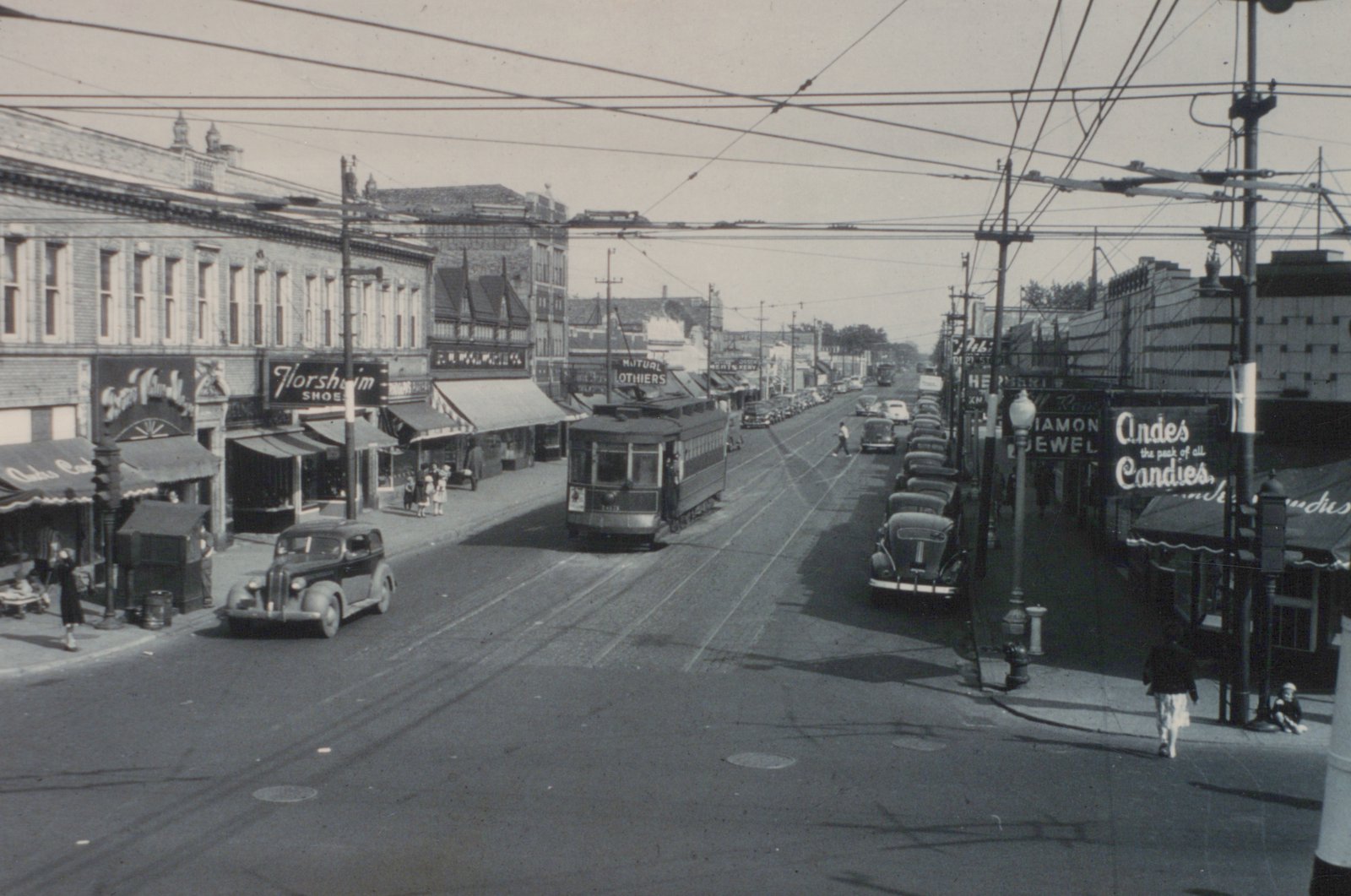 A black and white photo of a vintage streetcar on a city street, surrounded by parked cars and people on the sidewalks.