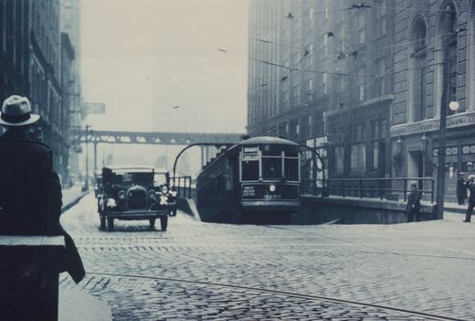 A black and white street scene from the 1930s showing a Chicago Surface Lines streetcar on a cobblestone street with an elevated railway in the background.