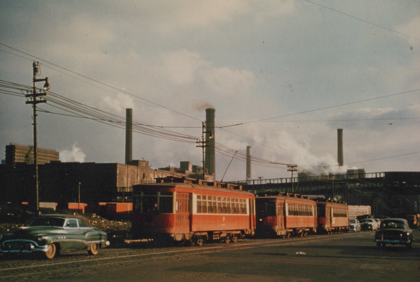 A line of red interurban railcars on a city street with steam rising from factory chimneys behind them, surrounded by 1950s cars.