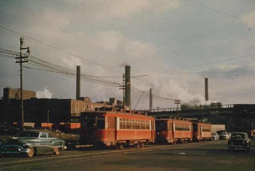 A line of red interurban railcars on a city street with steam rising from factory chimneys behind them, surrounded by 1950s cars.