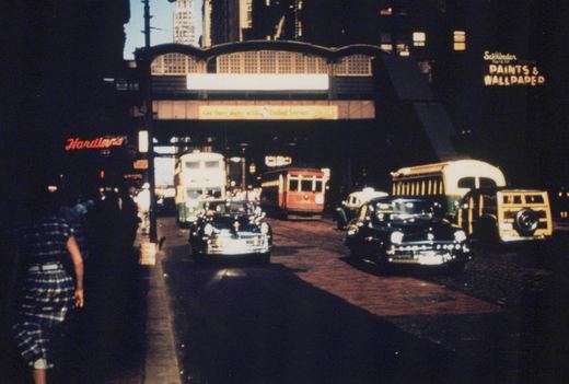A busy city street at dusk with vintage cars, a red trolleybus, and green-and-cream double-decker buses under an elevated train structure, with neon signs for 'Hardin's' and 'Schroder Paints' in the background.