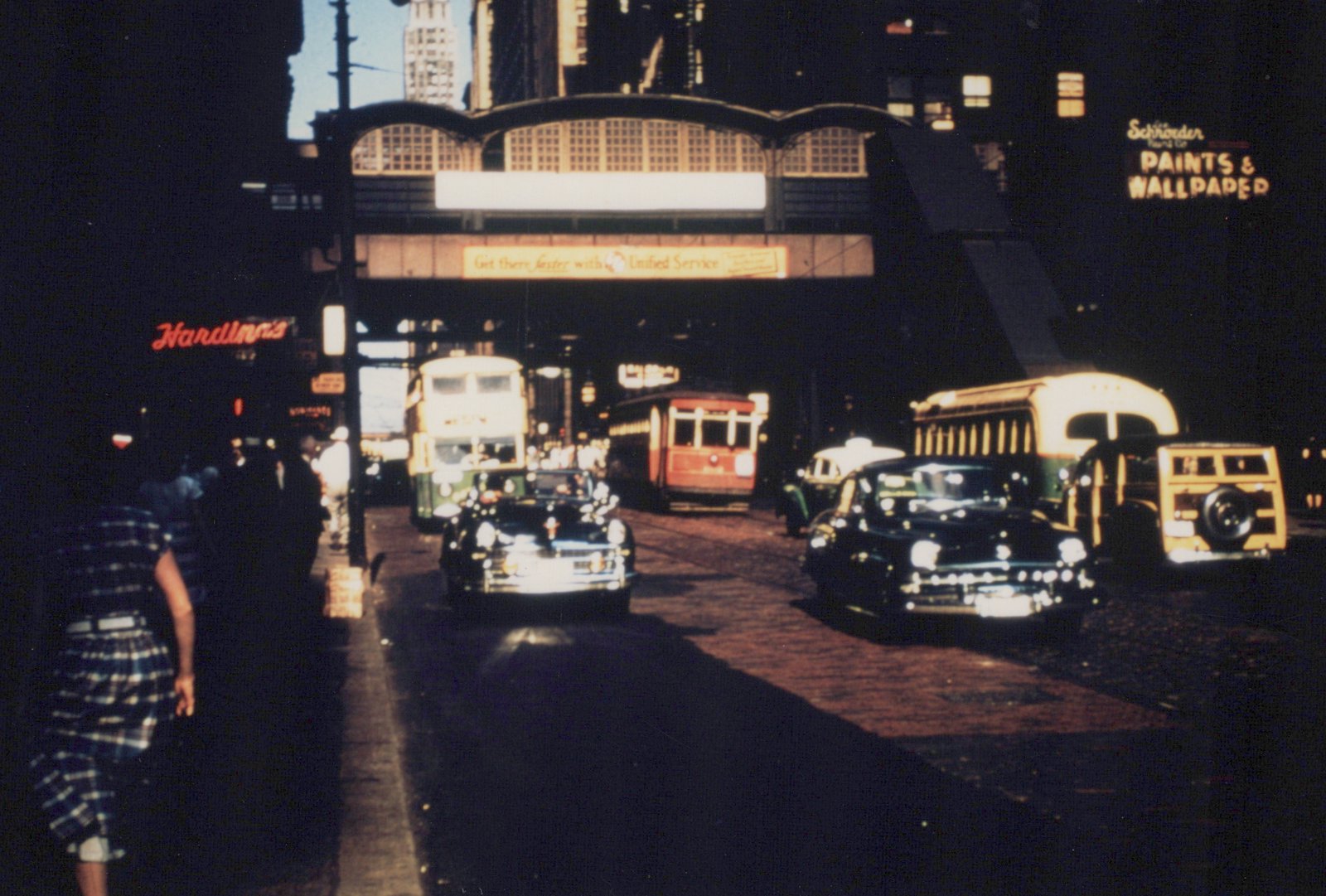 A busy city street at dusk with vintage cars, a red trolleybus, and green-and-cream double-decker buses under an elevated train structure, with neon signs for 'Hardin's' and 'Schroder Paints' in the background.