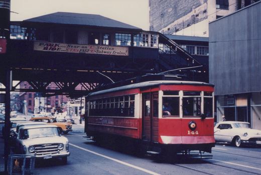 A red and white streetcar numbered 144 moves along a city street under a dark elevated train structure, with vintage cars and a billboard visible in the background.