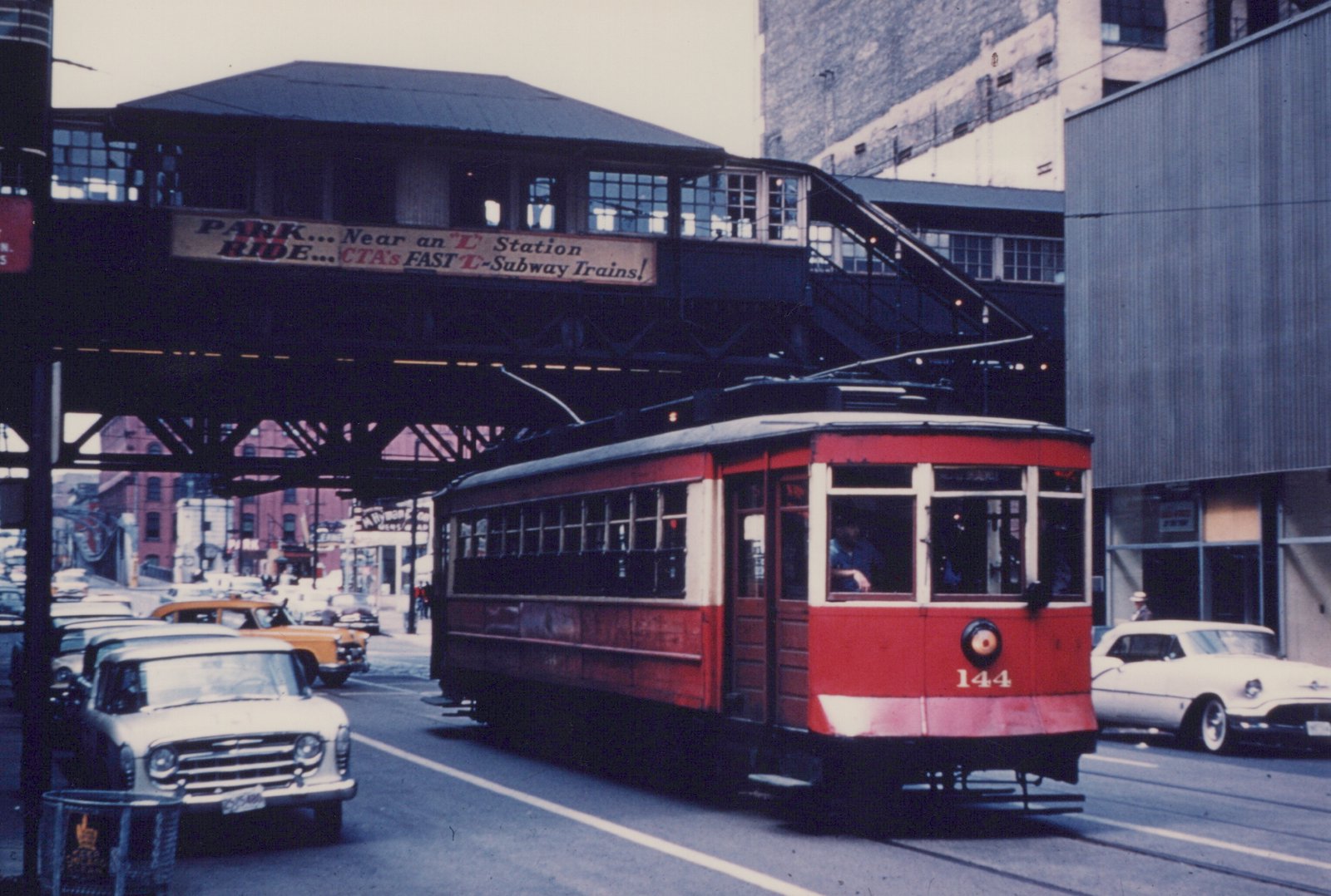 A red and white streetcar numbered 144 moves along a city street under a dark elevated train structure, with vintage cars and a billboard visible in the background.