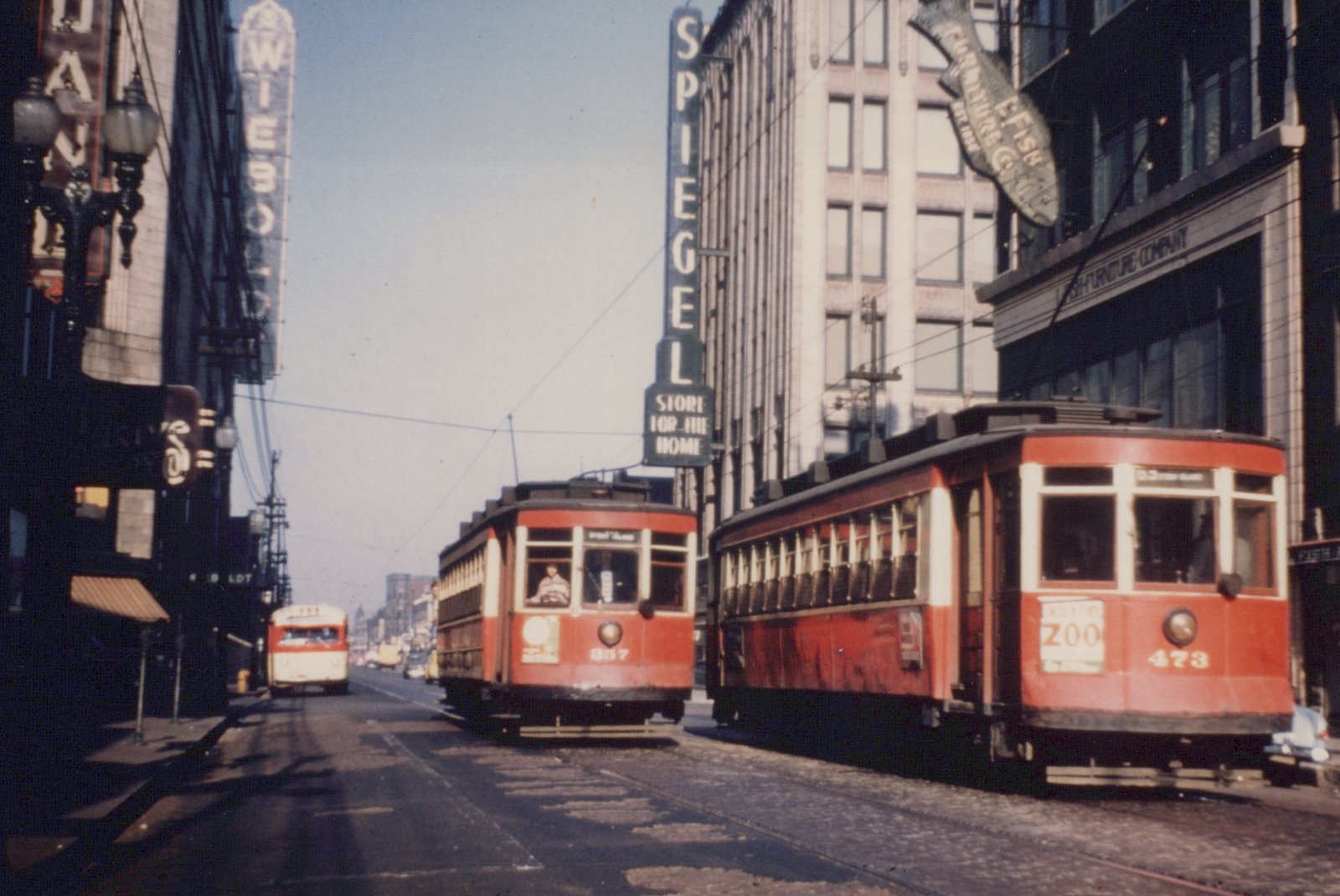 Two red and cream streetcars, numbered 473 and 857, are stopped on a city street lined with tall buildings and large store signs for Spiegel and Wieboldt under a clear sky.