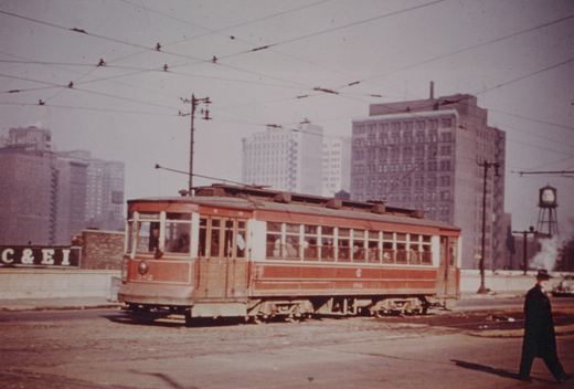 A vintage red and cream streetcar with the number 3152 on its side and front, running on city streets with tall buildings and a water tower in the background.