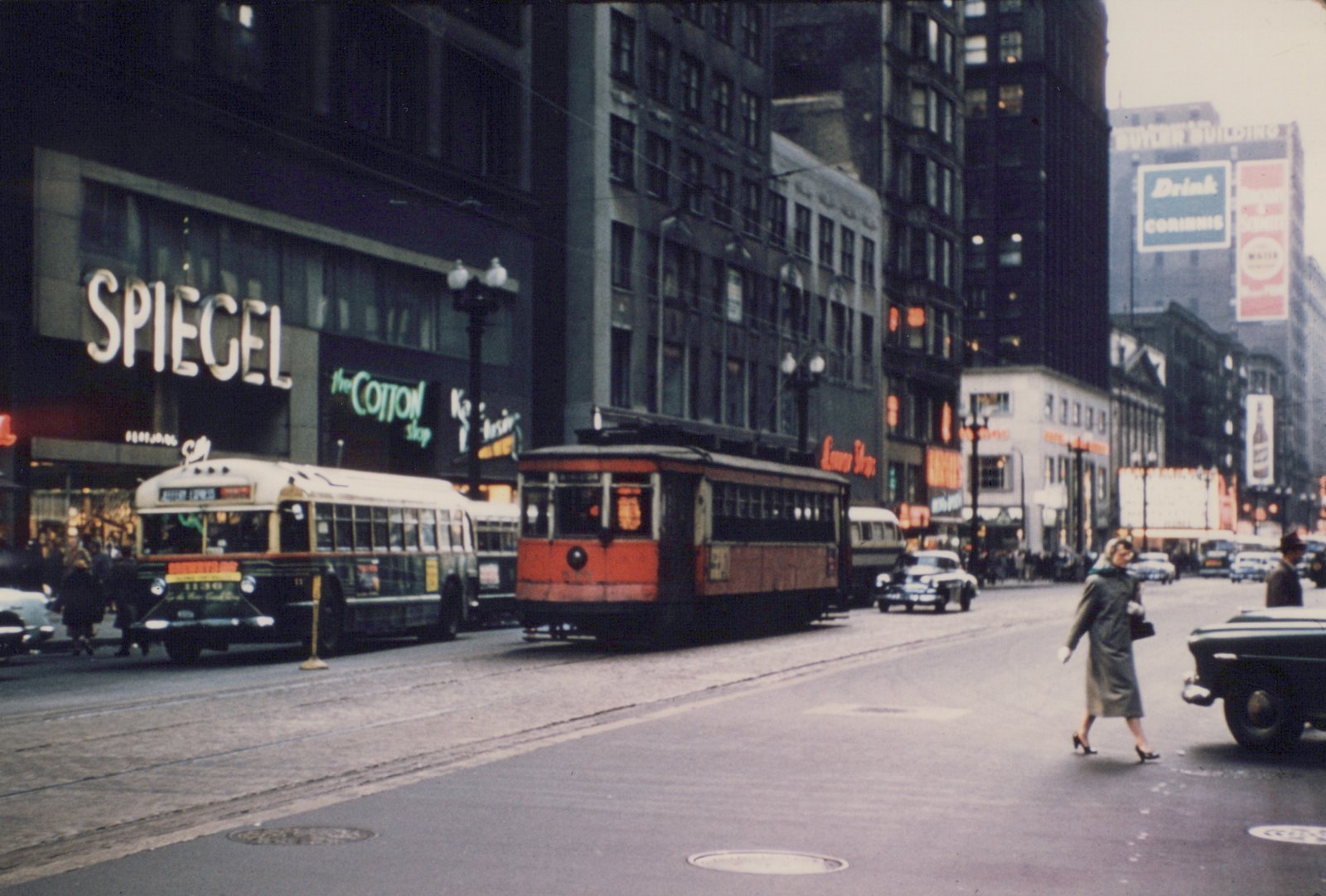 A red and green streetcar and a green and cream bus are on a city street with tall buildings, neon signs for 'SPIEGEL' and 'the COTTON shop', and mid-1950s cars. A woman in a coat is crossing the street.
