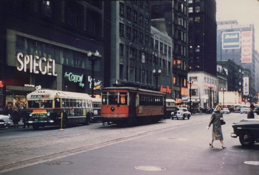 A red and green streetcar and a green and cream bus are on a city street with tall buildings, neon signs for 'SPIEGEL' and 'the COTTON shop', and mid-1950s cars. A woman in a coat is crossing the street.