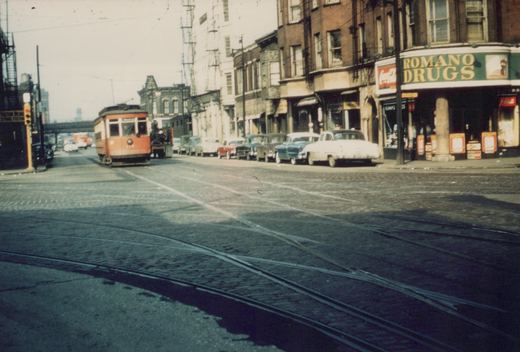 A red and cream streetcar with the number 317 on its front travels down a cobblestone street lined with 1950s cars and buildings with signs reading 'Romano Drugs' and 'Coca-Cola'.
